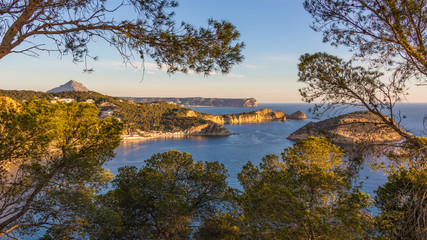 A wonderful view over the cliffs and bays of the northern Costa Blanca near the city of Javea in the sunshine and blue sky with trees and branches in the foreground.