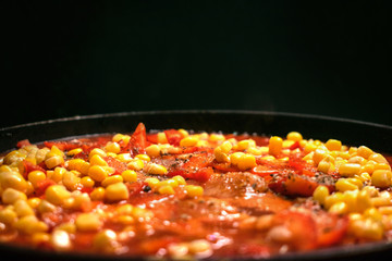 stewed vegetables, corn, tomatoes, sweet peppers, potatoes, miao in a pan on a dark background. Close up view