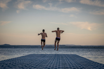 Young boys jumping in the sea from floating pier at sunset. First summer days. 