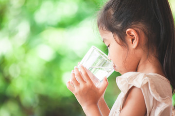 Cute asian child girl drinking fresh water from glass on green nature background