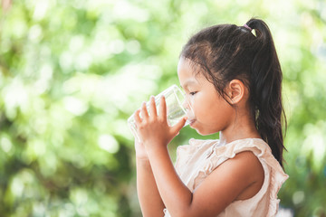 Cute asian child girl drinking fresh water from glass on green nature background