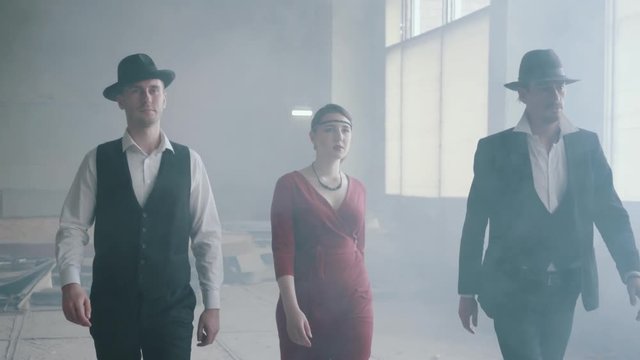 Two confident men in hats and suits and woman in red dress walking forward an abandoned building. The mafia in an empty building. Cool guys, thug, mafia, criminal gang, familia. Slow motion.