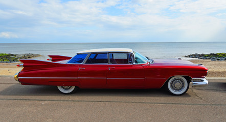 Classic Red 1950's 4 door Cadillac  motor car parked on seafront promenade. - obrazy, fototapety, plakaty