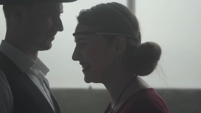 Handsome confident man in a hat whispering joke on the ear of elegant lady, she is laughing. The head of the mafia with his young lovely wife. Criminal authority, cool guys, mafia