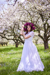 Obraz na płótnie Canvas Young beautiful girl in a wreath of lilacs. White lace dress on the perfect figure of the model. Photo session in the spring apple orchard in the open.