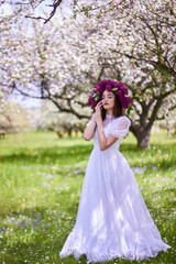 Obraz na płótnie Canvas Young beautiful girl in a wreath of lilacs. White lace dress on the perfect figure of the model. Photo session in the spring apple orchard in the open.