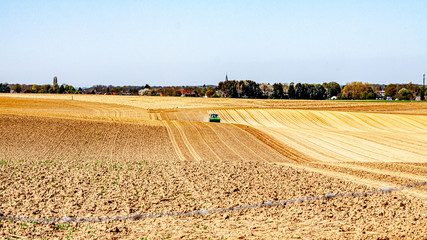 Fototapeta premium Agricultural farmland with a tractor plowing land with trees and villages in background against blue sky, preparing land to grow potatoes, sunny day in Oensel South Limburg, Netherlands