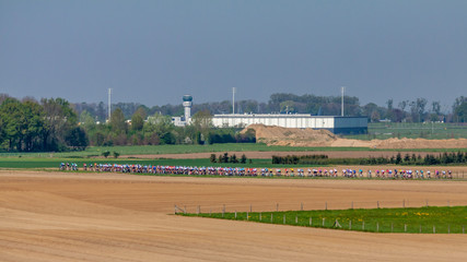 Fototapeta na wymiar Group of cyclists pedaling on a road in a cycling competition between farm fields and an airport in the background, wonderful spring day in the countryside in south Limburg in the Netherlands