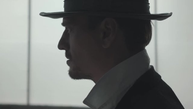 Portrait of handsome confident man putting a hat on the head in front of big window. The head of the mafia preparing for meeting. Criminal authority portrait. Self-confident businessman relaxing