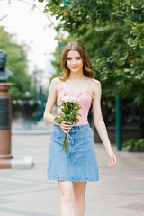 Portrait of a beautiful girl walking in the city, young smiling woman with a bouquet of flowers from pink and white roses, summer city background