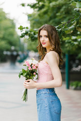 young beautiful woman in a pink top and blue denim skirt, a bouquet of flowers, pink and white roses walks along a city street in a public garden