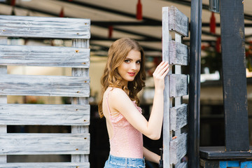 A beautiful young girl with flowing hair in a pink top and a blue skirt is standing near a wooden old gate, holds her hand on the gate and smiles. Summer day