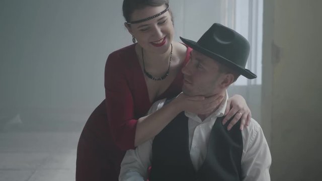 Handsome confident mafia boss in a hat and a vest sitting in an abandoned building. The elegant woman in red dress coming from behind and hugging the man smiling. Happy couple of gangsters