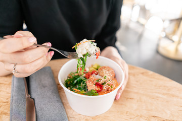 Woman eating traditional Hawaiian dish poke bowl with fork. Shrimps with rice, radish,cucumber,...