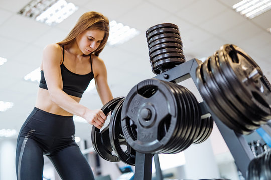 Concentrated young female in black sporty wear taking barbell disk in the gym. Beautiful fitness lady puts barbell plate into thespecial stand after healthy workout.
