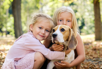 Little smiling blond girls sitting together hugging beagle dog in a sunshine autumn park