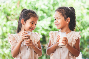 Two cute asian child girls drinking a milk from glass together on green nature background