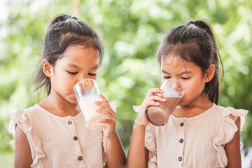 Two cute asian child girls drinking a milk from glass together on green nature background