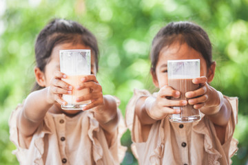 Two cute asian child girls drinking a milk from glass together on green nature background