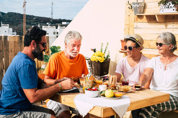 Adult people and one teenager smiling and eating. Multi generation family enjoy breakfast together. Senior couple with son and nephew. Wooden table and background