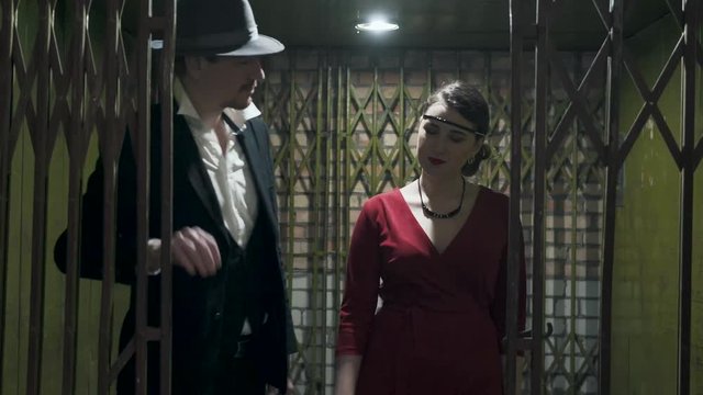 Portrait tall man in the suit and hat opens the door and passes inside the dark room a confident beautiful girl in an elegant red dress. Retro, vintage style. Mafia life