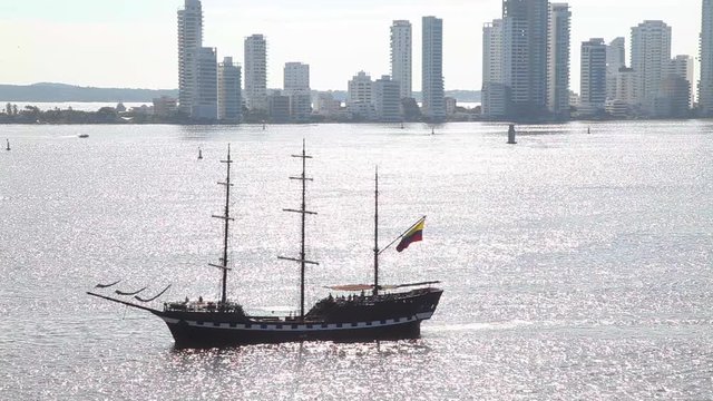 Old wooden Schooner with Columbian flag cruises in the bay with sun reflecting off the water and buildings visible in the background