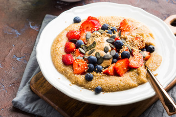 Delicious amaranth topped with almond butter, fresh strawberries and blueberry, sesame, pumpkin seeds. Plant based breakfast served on dark wooden board. Healthy colorful gluten free food concept.