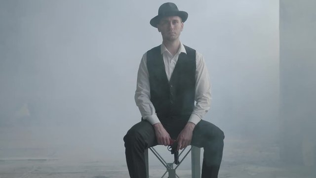 Handsome confident well-dressed man in a hat sitting on the chair holding a gun in the smoky room in an abandoned building. The head of the mafia is in an empty building. Cool guy, thug, mafia