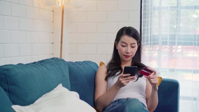 Young smiling Asian woman using smartphone buying online shopping by credit card while lying on sofa when relax in living room at home. Lifestyle women at house concept.