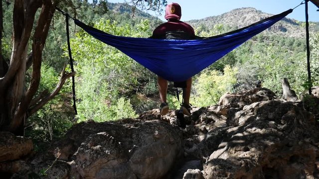 Guy wearing hat sitting in a blue hammock stretched between two short trees rocking back and forth looking into the distance with mountains and green trees in the background in slow motion.