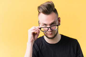Portrait of sceptic millennial guy on yellow background. Young emotional man taking of glasses with judgy facial expression.