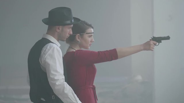Elegant young woman in red dress slowly aiming a gun, handsome gallant man stands behind, directing her hand, side view. Happy couple of gangsters. Bonnie and Clyde. Side view