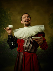Young man as a medieval knight on dark studio background. Portrait in low key of male model in retro costume. Drinking morning coffee. Human emotions, comparison of eras and facial expressions concept - obrazy, fototapety, plakaty
