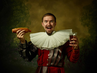 Young man as a medieval knight on dark studio background. Portrait of male model in retro costume. Holding a glass of beer and hot-dog. Human emotions, comparison of eras, facial expressions concept. - obrazy, fototapety, plakaty