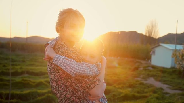 Beutiful young blonde mother hugging her child at green field against a golden sunset , the concept of a happy family and leisure time spending during warm summer holiday, son dressed in cage shirt
