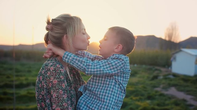Adorable young mother kissing her charming child at green field with gold warm sunset, happy weekend time spending together, content of enjoyable family leisure lifestyle at countryside of Spain