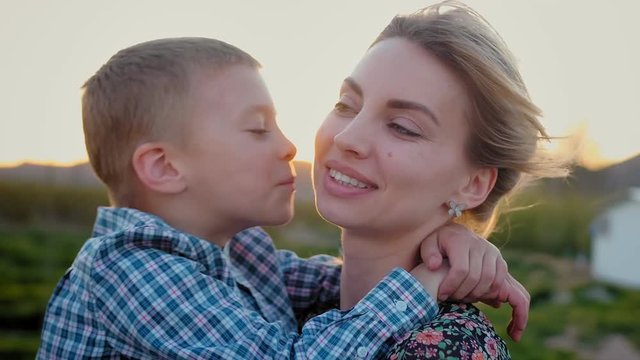 Portrait of pretty blonde small boy kissing and hugging his charming mother at countryside green field, enjoying spending adventure positive time together at fresh nature, child dressed in cage shirt