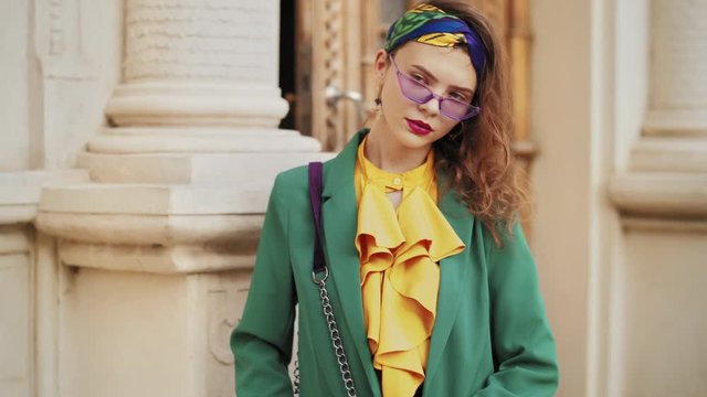 Street fashion portrait of young beautiful elegant lady wearing trendy purple sunglasses, headband, stylish yellow blouse, green blazer, with quilted shoulder bag, posing in old european city