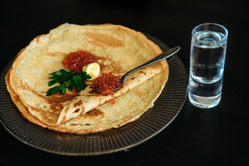 Golden russian pancakes with red caviar and butter in transparent plate near the glass of ice vodka isolated on black background. Restaurant of national cuisine.