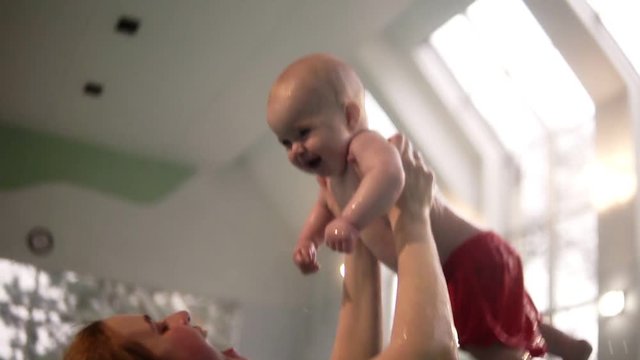 Mother and her baby todler in cute red shorts having fun together in the indoors swimming pool. Playing in the water. Healthy family lifestyle and children water sports activity. Slow motion