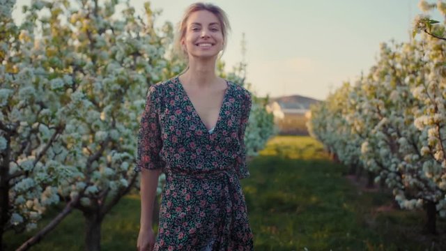 Happy trendy blonde girl goes bouncing through white spring blossoming garden at Europe, showing positive vibe and pleasant emotional content of free vacation time, dressed in colorful flower print
