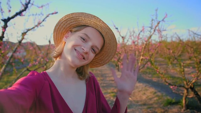 Portrait of beautiful blonde girl smiling online to friend call on mobile phone at flower fields, dressed in straw summer hat and beautiful summer pink dress, cheerful trendy model vloger using video