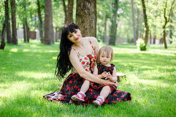 Mother and little lovely daughter sits on green grass under trees shades at sunny summer park. Childhood and motherhood. Concept of a happy family. Mother and kid. Universal Children's Day