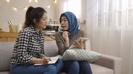 Two happy multi ethnic female students studying checking and comparing notes sitting at home...