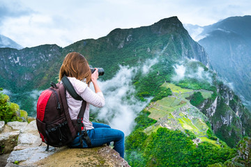 Girl with hiking rucksack sitting on the edge of the rock and taking amazing photos of breathtaking Machupicchu mountains landscape