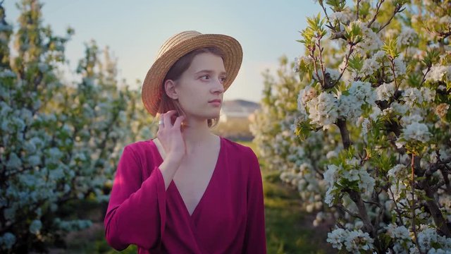 Portrait of sensible young female gently touching her blonde hair covered with hat outdoor, lonely trendy calm girl standing between blossoming colourful farm bushes at sunny countryside evening light