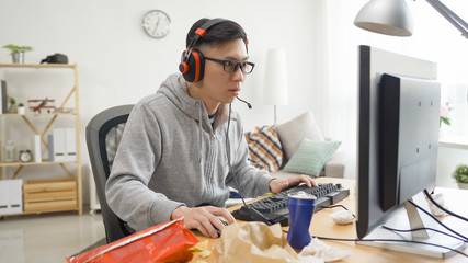copy space young asian man playing shooting video game inside messy room at home. high school senior teenage boy student using computer having fun wear headphones with junk food and snack chips.