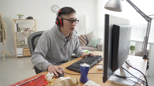 Close up of young asian man in headphones playing computer game and being angry as losing mistake. male college student sitting in bedroom having fun on pc mad beat bang on keyboard with fist.