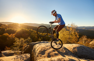 Fototapeta na wymiar Young cyclist standing on back wheel on trial bicycle. Sportsman rider making acrobatic stunt on the edge of big boulder on the top of mountain at sunset. Concept of extreme sport active lifestyle