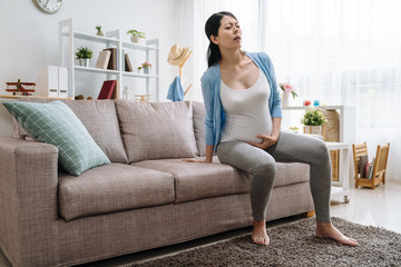 Obraz premium illness pregnant asian woman mom sitting down with difficult. young girl future mother hands holding heavy belly with boy baby inside prepared to resting on sofa couch hard to move heavy body at home